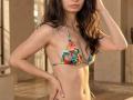 VikkiRi in a colorful bikini with tropical patterns, showing her small tits and slim, tan, flat-stomached body, long dark hair, and hand touching her head.