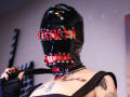 AmySpider with black latex mask featuring red and black metal rings, chain collar, tattooed chest, black sleeveless top, holding black leather strap, black nail polish, in a room.