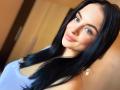 Olieri has black, shiny hair, blue eyes, and a cute smile. She's wearing a purple top, showing her small tits, with a subtle blush on her fair skin.