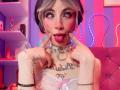 LaurenEvie69 with tongue out, sticking fingers near mouth, wearing white lace top, pink bow, silver choker, and tattoos, in a neon-lit room with pink and purple lights.