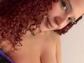 ValeryScot with curly red hair, light skin, and a nose ring, smiling at the camera, showing her bare tits, wearing a necklace with a green gem, in a selfie.