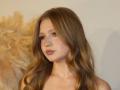 AkilahGhil has long, wavy brown hair, light skin, and is looking to the side with pink lipstick and subtle eye makeup. She's wearing a feathered accessory on her shoulder.