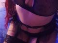 AntoniaBrunni wearing black mesh lingerie, showing her tits and pussy through the fabric. She has red hair, lace gloves, and a black belt. Blue bed in background.