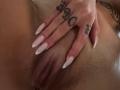 AuraXo's hand with long, pale pink nails rests on her wet, brown-skinned pussy, showing tattooed fingers. Her vulva is visible, slightly parted.