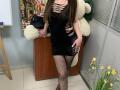 Omella with long brown hair wearing a black, torn, mesh dress, fishnet stockings, and heels. She's holding a white teddy bear, standing in a room with yellow flowers.