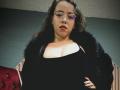 BarbaraLeythonn with glasses, dark wavy hair, red lipstick, black fur coat, low-cut black top showing her big tits, standing, hand on hip, serious look, red tufted chair.