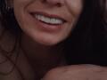 SkylerSamuel's face with a smile, showing white teeth, wearing headphones, with brown hair, and a visible shoulder in a dimly lit, dark photograph.