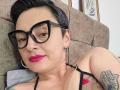 KatParadise con capelli neri corti, occhiali neri, rossetto rosa, piercing al naso, orecchini a cerchio, bretella nera, collana di perle, tatuaggio sul braccio, sdraiata su un divano marrone, che guarda la fotocamera..