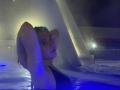 KrystalBerkovitch, with wet blonde hair, arms raised, in a dark blue-lit pool, wearing a black one-piece, looking slightly annoyed, with blue water and a misty background.