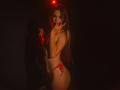 AgataRyili with long brown hair, red string bikini, and tattoos, standing in dim light, covering her mouth with her fingers, red light above her head, showing her ass.