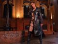 VanasValet in black leather coat, strappy lingerie, and thigh-high boots, standing in a dimly lit brick building, with her hand on her head, showing her ass.