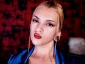 FiorellaNox has blonde hair, blue eyes, and red lipstick. She's wearing a blue satin shirt, black ear gauges, and a thin gold necklace. Her tongue is slightly out.