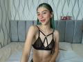 AbbyHillsI with green-blue hair, small tits in black sheer bra, sitting on bed, smiling, fair skin, ear gauges, cute, bedroom setting, bed with grey headboard.