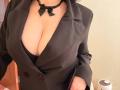 EmmaNoa indossa un blazer nero, una collana con un fiocco, mostrando il suo seno abbondante in un reggiseno nero. La sua mano è appoggiata sul fianco, mettendo in risalto le sue curve..