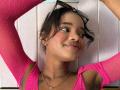 KiaraBrown with dark hair, pink sparkly top, arms raised, eyeglasses on head, smiling, tan skin, small tattoo on left arm, gold necklace, brown eyes, slight blush on cheeks.