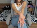 Rozalin, sitting in a grey chair, wearing a white tank top and blue jeans, with hands resting on her knee, choker necklace, brown hair, small tits, wooden floor.