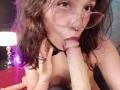 SophieBlake with wavy brown hair, nose ring, and glasses, is fellating a large, erect, circumcised dick. Her hand is on her tit, and her tongue is wrapped around it.