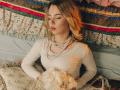 KylaHemeke with wavy blonde hair, wearing a white lace dress, pearl necklace, holding a bouquet of white flowers, eyes closed, sitting on a floral-patterned bed.