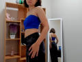Steisii with blue strapless top, showing small tits, wearing black jeans, reflected in mirror. Her ass is visible, and she has a necklace. Room has wooden shelves and plants.