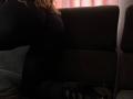 Niferttiti with large tits, wearing a tight black top and black pants, kneeling on a dark gray couch, with curly blonde hair and black high heels in the foreground.