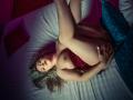 VeraNoires is lying on a bed, legs up, wearing a red satin skirt, exposing her tits, with blonde hair and red lipstick, holding her pussy, in a dark room.