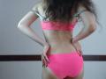 VikkiRi in a pink bikini, back view, hands on hips, showing a slim, toned ass, small tits, long wavy brown hair, black nail polish, gray strap, pink bottoms.