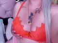 AlessadraJoness con un reggiseno di pizzo arancione, una mano sulle sue grandi tette, tatuaggi sul petto, lunghi capelli argentati, una collana con un cuore, in una stanza rosa con decorazioni a forma di ciuffi..