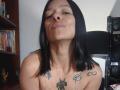 AmelieBlake with black hair, nose ring, and tattoos, smiling with puffed cheeks, topless, showing her tits and tattoos, sitting in a room with bookshelves.