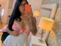 CatalinaReiyna with glasses, black hair, tattooed arm, large boobs in sheer pink and white lingerie, holding orange phone, mirror selfie in bedroom with lamps.