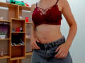 Steisii with red lace bralette showing small tits, belly button, and hands in blue jeans. Her head is out of frame. She's standing in a room with wooden shelves.