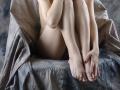 KamiLove's pale legs and feet are crossed, with hands gently touching toes, sitting on a chair covered in crumpled, grayish fabric, showing her pink-nailbed toes.