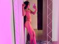 MilanMoon, nude with black hair, leans against a glowing pink wall, holding a phone in a mirror selfie. Pink lights highlight her slim, sexy body, small breasts, and confident stance.