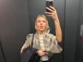 ArnettaNazario with blonde hair, tongue out, taking a mirror selfie in a dark elevator, wearing a white shirt, gray patterned jacket, holding a black bag, red nails.