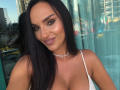 CrazyDeea69 with long black hair, brown eyes, and a smile, wearing a white lace top, showing her tits, and a silver necklace, standing in front of a glass window.