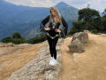 EliseMonet, blonde, standing on rocky ledge, wearing black dress with white floral patterns, black leggings, white sneakers, hands on hips, mountains and trees in background, looking at camera.