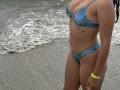 Sarah, une princesse en bikini bleu avec des gouttes d'eau, montrant ses seins et ses fesses, a un tatouage sous ses seins, porte un bracelet jaune et se tient près de l'océan..