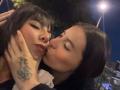 Two women with black hair, one with a tattoo on her hand, kissing passionately at night. The one on the right is kissing the left one's lips, eyes closed. Streetlights in background.