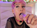 DannyRouds with pink hair, black mesh top, tattooed arm, pink choker, and ear piercings, looking at camera with a slight smile. Soft purple and white lighting.