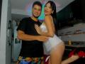 FexCouplee69 with long black hair, red lipstick, white sheer top, and red high heels, leans into a bearded man in a black t-shirt and colorful shorts, kitchen background. Sexual tension.