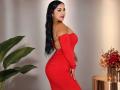 LanaRossel with long black hair, wearing a tight red off-shoulder dress, showing her ass and small tits, standing beside a plant and a flower vase.