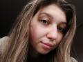 Sanella's face, with her brown eyes, pink lips, and slightly flushed cheeks, has long, wavy brown hair and a subtle smile. Her nose is small and she's looking at the camera.