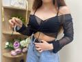 Rioria with small tits, wearing a black sheer off-shoulder top and blue denim skirt, holding black strings, standing in front of a wooden shelf with flowers.