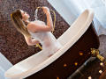 Karolichee in a white, wet, see-through dress, standing in a brown and white vintage bathtub, holding water from a gold faucet, with candles on the floor.
