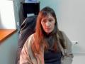 MiraMixi has long brown hair, a nose piercing, and wears a beige jacket over a black shirt. She sits in a black office chair, looking at the camera.