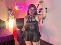 LilithDark69 with purple hair, black lipstick, and glasses, holding a leather strap, wearing a black leather dress with buckle details, showing tattoos, standing in a dimly lit room.