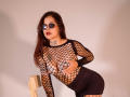 ElectraRoss with long brown hair, wearing reflective sunglasses, black fishnet top exposing her tits, black skirt, and wooden chair, leaning forward, red lipstick, tattoo on neck.