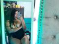 AriaVega with wet curly hair, topless, covering her tits with a black one-piece, taking a mirror selfie, showing her ass and a teasing look, in a green-lit room.