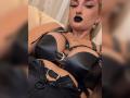 MademoiselleBella with black leather harness, showing her large tits, black lipstick, and earrings, lying back, wearing high-waisted black leather pants, with metal buckles and straps.
