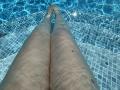 Barbarica's legs in a pool, water up to mid-thigh, blue-tiled floor, sun reflecting, legs slightly apart, skin tone is light, small dark spot on right thigh.
