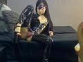 MiaDeLune, vêtue d'une combinaison noire brillante en latex, avec des bottes montantes et des culottes roses, montrant sa vulve, prend un selfie avec son téléphone, sur un lit noir..